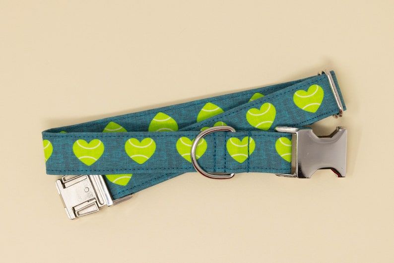 May include: A teal dog collar with a repeating pattern of lime green hearts. The collar has a silver metal buckle and a D-ring for leash attachment. The background is a solid beige color.