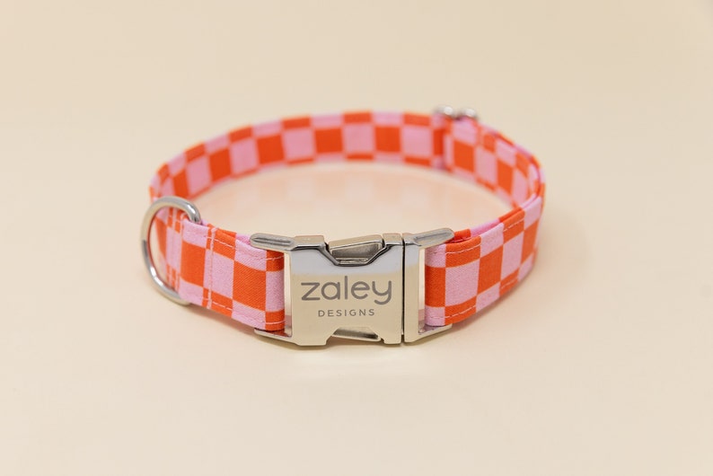 Op de afbeelding: Een roze en oranje geruite hondenriem met een zilveren gesp waarop "zaley designs" staat.
