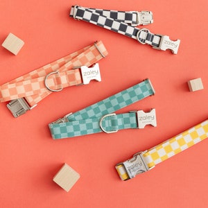 May include: Four dog collars with a checkered pattern in different colors: black and white, peach, teal, and yellow. The collars have silver buckles with the word "Zaley" printed on them.