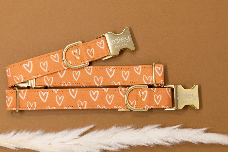 May include: A gold dog collar with a white heart pattern on a peach background. The collar has a gold buckle with the text "zaley designs" on it.