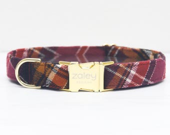 May include: A plaid dog collar in shades of burgundy, orange, and brown. The collar features a gold-colored metal buckle and a D-ring. The text "zaley DESIGNS" is printed on the buckle.