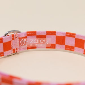Op de afbeelding: Oranje en roze geruite hondenriem met een zilveren gesp en een wit label met de tekst "zaley designs.com"