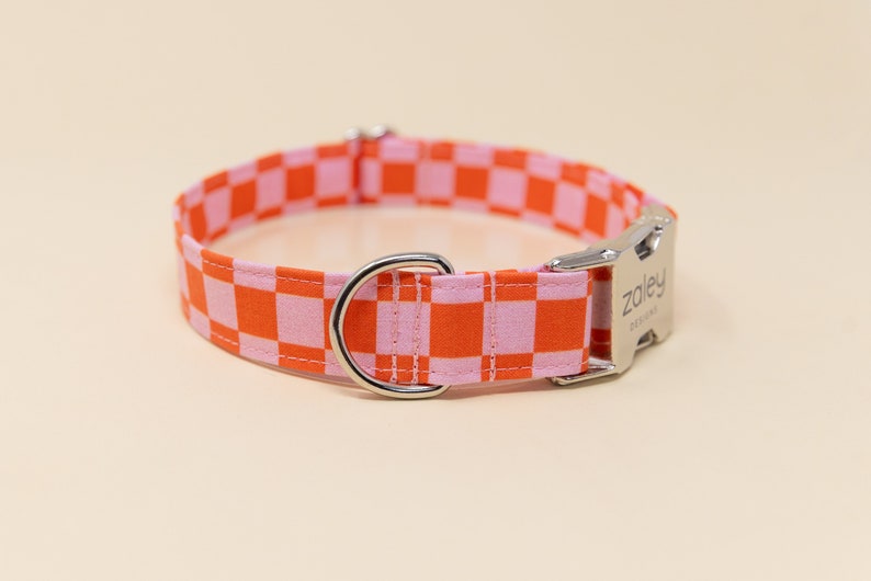 Op de afbeelding: Een roze en oranje geruite hondenriem met een zilveren gesp en een zilveren D-ring. De gesp heeft de tekst "Zoley Designs" gegraveerd.