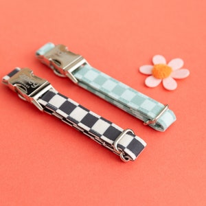 May include: Two dog collars with checkerboard patterns. One collar is black and white, the other is light blue and white. Both collars have silver buckles and are on a red background.