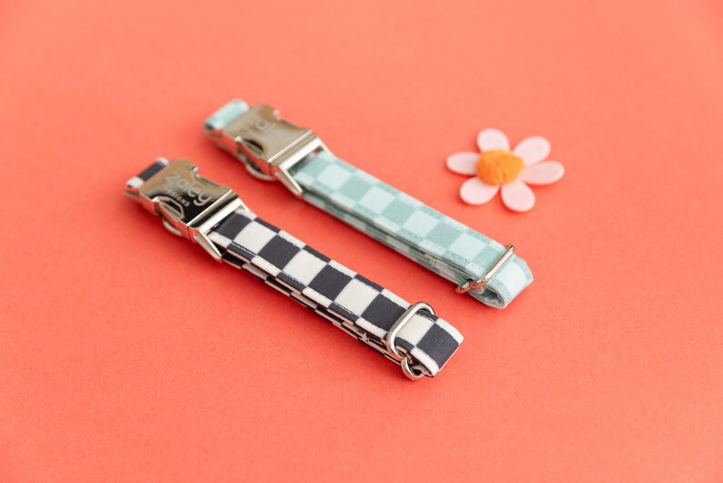 May include: Two dog collars with checkerboard patterns. One collar is black and white, the other is light blue and white. Both collars have silver buckles and are on a red background.