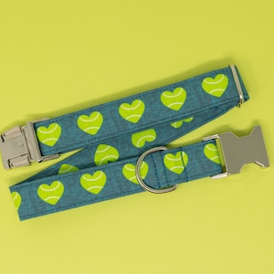 May include: A teal dog collar with a pattern of lime green tennis ball hearts. The collar has a silver metal buckle and a D-ring for leash attachment. The background is a solid yellow-green.