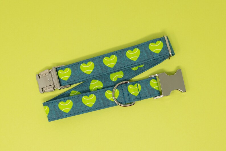 May include: A teal dog collar with a pattern of lime green tennis ball hearts. The collar has a silver metal buckle and a D-ring for leash attachment. The background is a solid yellow-green.