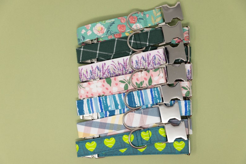 May include: A collection of dog collars with various fabric patterns. Designs include floral, plaid, lavender, and striped patterns. Each collar has a silver metal buckle and a D-ring for leash attachment. The collars are displayed on a light green background.