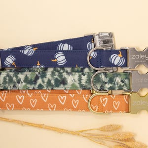 May include: Four dog collars with different patterns. The collars are stacked on top of each other. The top collar is navy blue with white pumpkins. The second collar is green with a forest pattern. The third collar is orange with white hearts. The collars all have silver buckles with the word "Zaley Designs" engraved on them.