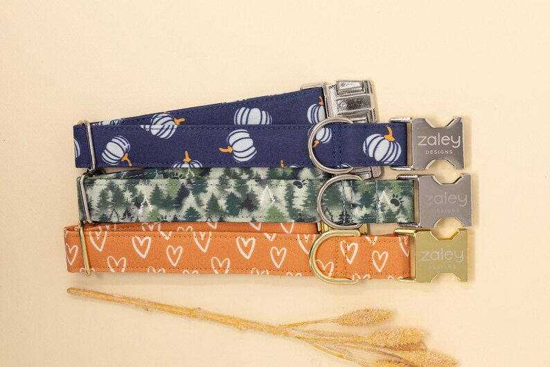 May include: Four dog collars with different patterns. The collars are stacked on top of each other. The top collar is navy blue with white pumpkins. The second collar is green with a forest pattern. The third collar is orange with white hearts. The collars all have silver buckles with the word "Zaley Designs" engraved on them.