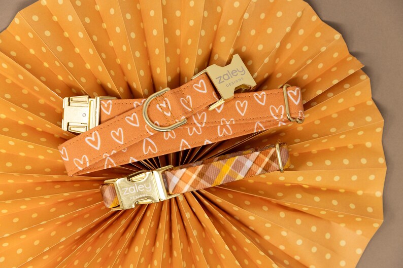May include: Two dog collars with gold hardware. One collar is orange with white hearts and the text "Zaley Designs". The other collar is orange, yellow, and brown plaid.