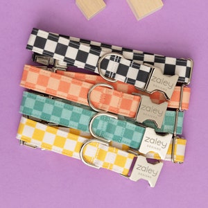 May include: Five colorful checkered dog collars with silver buckles. The collars are stacked on top of each other and feature the text "Zaley Designs" on the buckles.