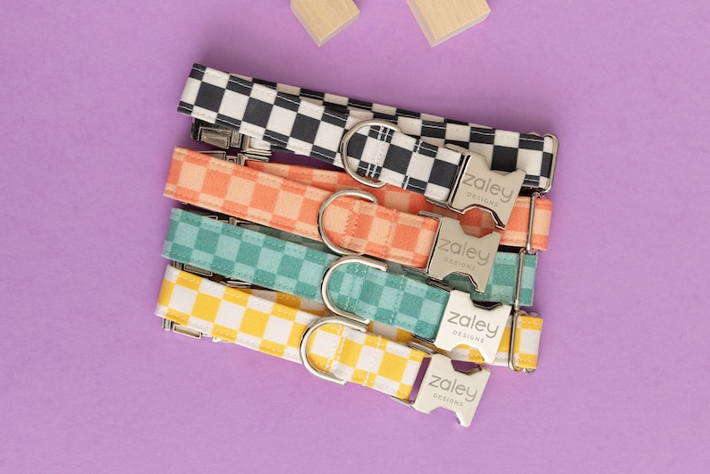 May include: Five colorful checkered dog collars with silver buckles. The collars are stacked on top of each other and feature the text "Zaley Designs" on the buckles.