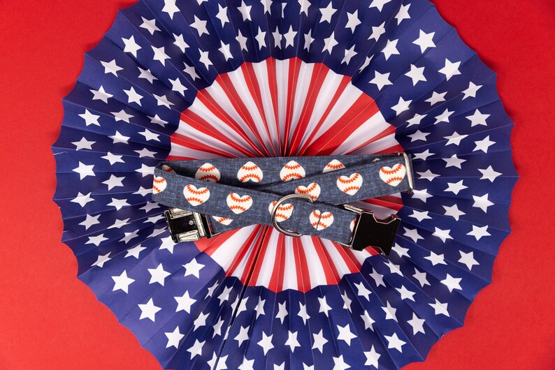 Puede incluir: Un collar de perro azul y blanco con un estampado de coraz&oacute;n rojo y b&eacute;isbol. El collar est&aacute; encima de un abanico de papel rojo, blanco y azul.