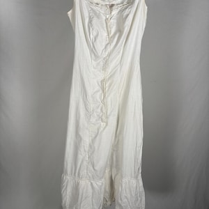 May include: A vintage white slip dress with a square neckline and delicate lace trim. The dress features a button-up front and a ruffled hem with eyelet detailing. It hangs on a wooden hanger against a plain background, showcasing its classic design.