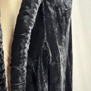 May include: A close-up of a black velvet garment, possibly a coat or jacket, draped on a mannequin. The fabric has a rich, textured appearance with visible folds and a slight sheen. The garment's design includes a padded collar and a slit detail.