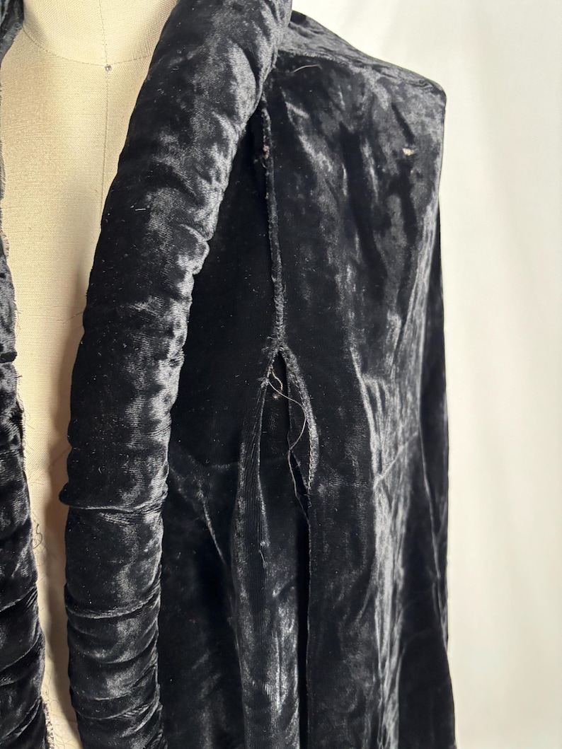 May include: A close-up of a black velvet garment, possibly a coat or jacket, draped on a mannequin. The fabric has a rich, textured appearance with visible folds and a slight sheen. The garment's design includes a padded collar and a slit detail.