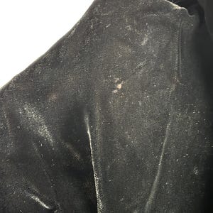 May include: Close-up of a section of black velvet fabric. The fabric has a soft, textured appearance with subtle variations in color and sheen. There are a few small, light-colored spots on the surface, adding a touch of visual interest to the dark material.
