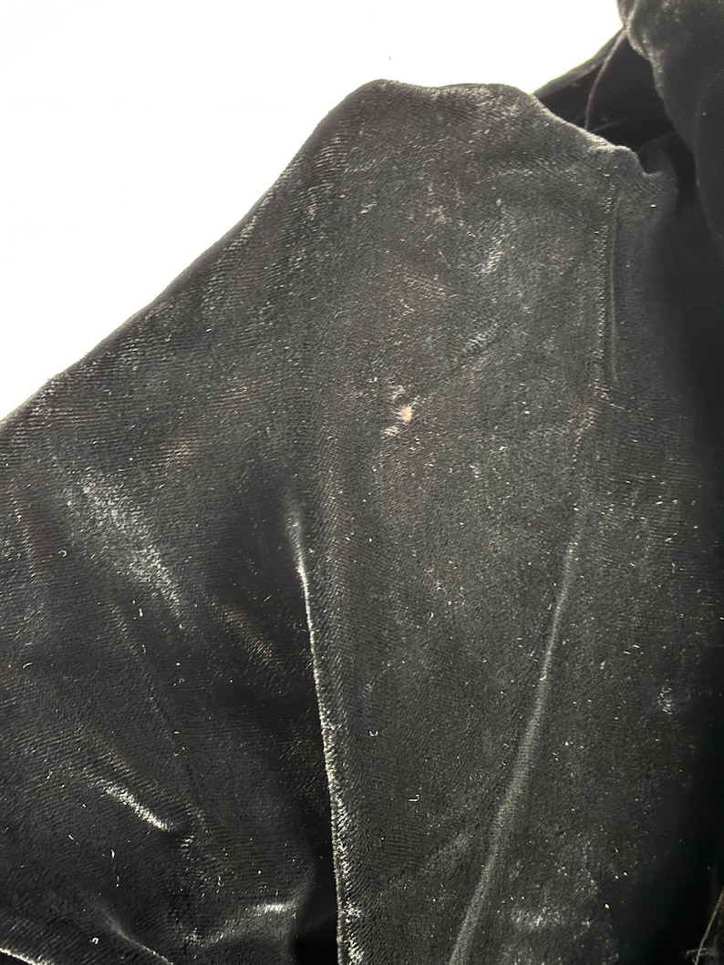 May include: Close-up of a section of black velvet fabric. The fabric has a soft, textured appearance with subtle variations in color and sheen. There are a few small, light-colored spots on the surface, adding a touch of visual interest to the dark material.
