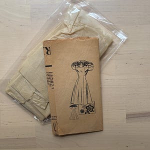 Könnte beinhalten: Vintage Schnittmuster mit einer Zeichnung eines Kleides auf der Vorderseite. Das Schnittmuster befindet sich in einer transparenten Plastiktüte, zusammen mit anderen Schnittteilen. Das Muster ist beige mit schwarzem Text und Illustrationen. Der Hintergrund ist helles Holz.