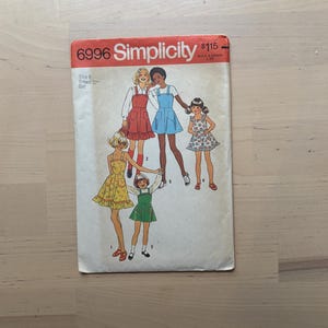May include: Vintage Simplicity sewing pattern 6996 for girls' dresses. The pattern envelope is red and white, featuring illustrations of the dress designs. Size 8, breast 27 inches. The Simplicity logo is prominently displayed.