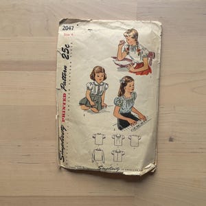 May include: A vintage Simplicity Pattern 2047, size 4, for children's clothing. The cover displays illustrations of girls in different outfits. The pattern's aged appearance suggests its vintage status, offering a nostalgic look at classic children's fashion.