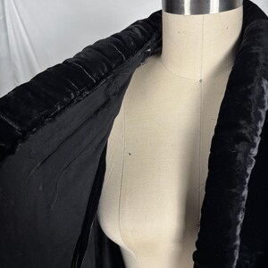May include: A black velvet coat collar is draped over a mannequin. The collar is thick and padded, with a textured, ribbed design. The interior lining of the coat is visible, revealing a smooth, black fabric. The coat is a vintage garment.