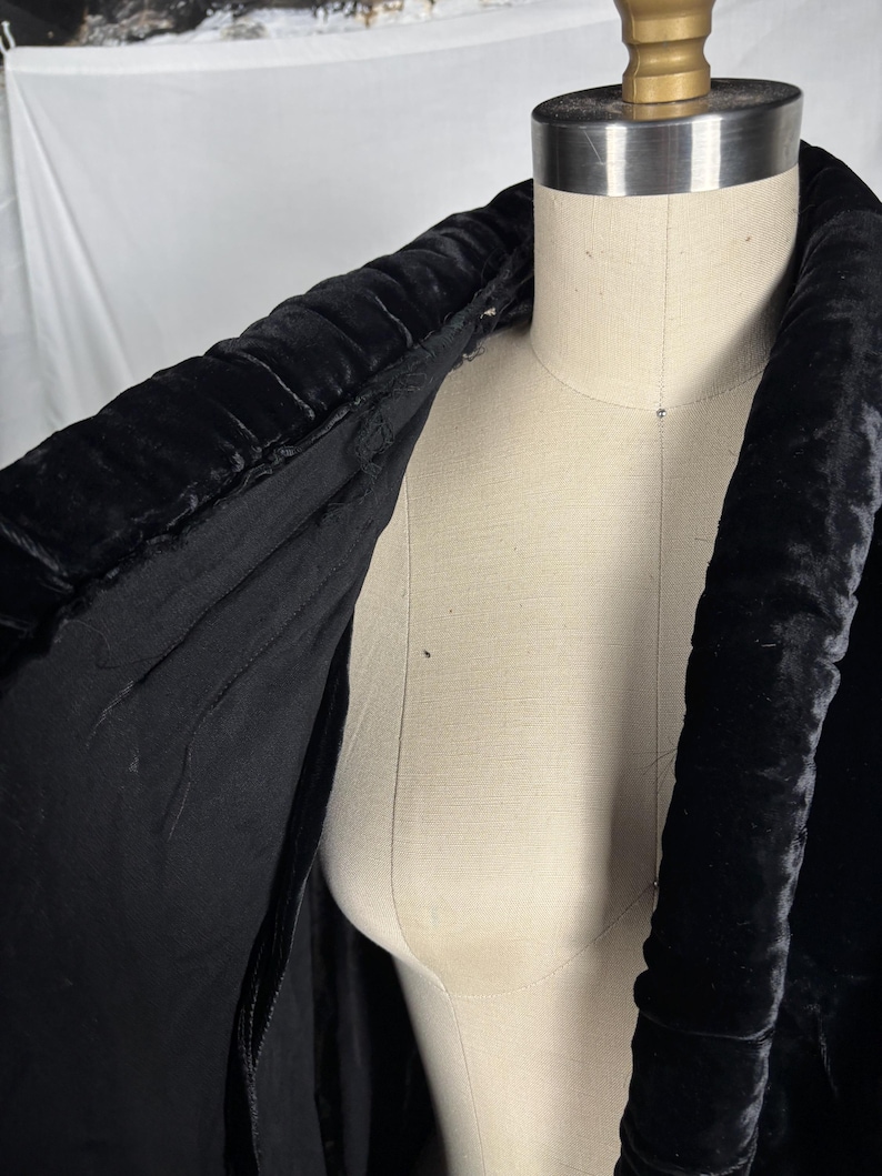 May include: A black velvet coat collar is draped over a mannequin. The collar is thick and padded, with a textured, ribbed design. The interior lining of the coat is visible, revealing a smooth, black fabric. The coat is a vintage garment.