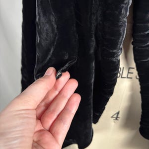 May include: Close-up of a black velvet jacket. The jacket has a button closure and a textured, ruched design. The fabric appears soft and luxurious, with a subtle sheen. The sleeves are long and gathered.