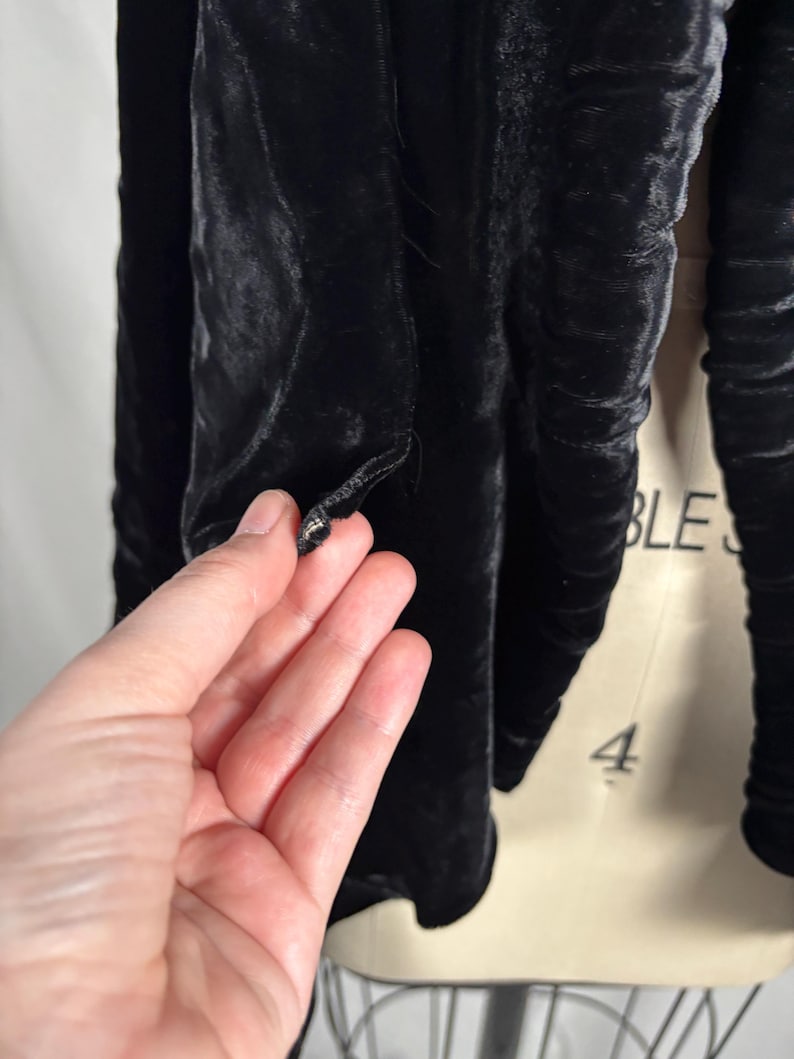 May include: Close-up of a black velvet jacket. The jacket has a button closure and a textured, ruched design. The fabric appears soft and luxurious, with a subtle sheen. The sleeves are long and gathered.