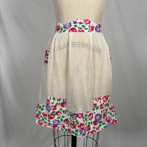 May include: A vintage white apron with a floral pattern. The apron features a dotted fabric with floral trim at the waist and hem. The trim includes red, pink, and purple flowers with green leaves. The apron is displayed on a mannequin.