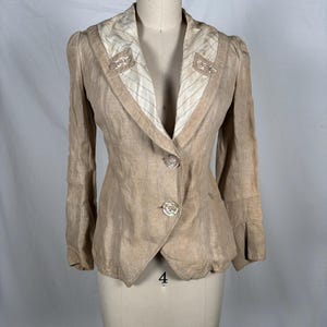 May include: A tan linen blazer with a notched lapel and two clear buttons. The blazer has a tailored fit, long sleeves, and decorative embellishments on the collar. The lining is striped.