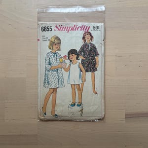 May include: A vintage Simplicity sewing pattern for girls' dresses, size 10, with a breast measurement of 28 inches. The pattern cover features illustrations of three girls wearing the dresses. The pattern is in a clear plastic sleeve.