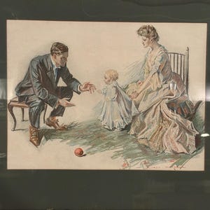 Puede incluir: Impresión vintage enmarcada que representa una escena familiar. Un hombre con traje y una mujer con vestido están con un bebé. La obra de arte está en un marco de madera oscura. La impresión tiene una paleta de colores suaves con una pelota en el suelo.