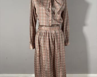 May include: A vintage two-piece dress set in a plaid pattern. The set includes a long-sleeved blouse with a large bow at the neck and a pleated midi skirt. The colors are primarily brown, pink, and white. The dress is displayed on a mannequin.