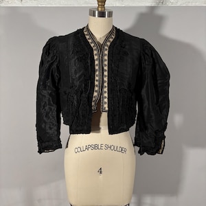 Puede incluir: Chaqueta corta negra con adornos decorativos y mangas abullonadas. La chaqueta se exhibe en un maniquí. El texto "COLLAPSIBLE SHOULDER" y el número "4" están impresos en el maniquí.
