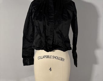 May include: A black, long-sleeved blouse with a high collar, displayed on a mannequin. The blouse has a slightly shiny texture and a button-up front. The mannequin has the words "COLLAPSIBLE SHOULDER" and the number "4" printed on it.