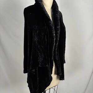 May include: A black velvet jacket with a draped, oversized collar. The jacket is displayed on a mannequin, showcasing its flowing design and soft texture. The garment has long sleeves and a relaxed fit, suitable for layering.