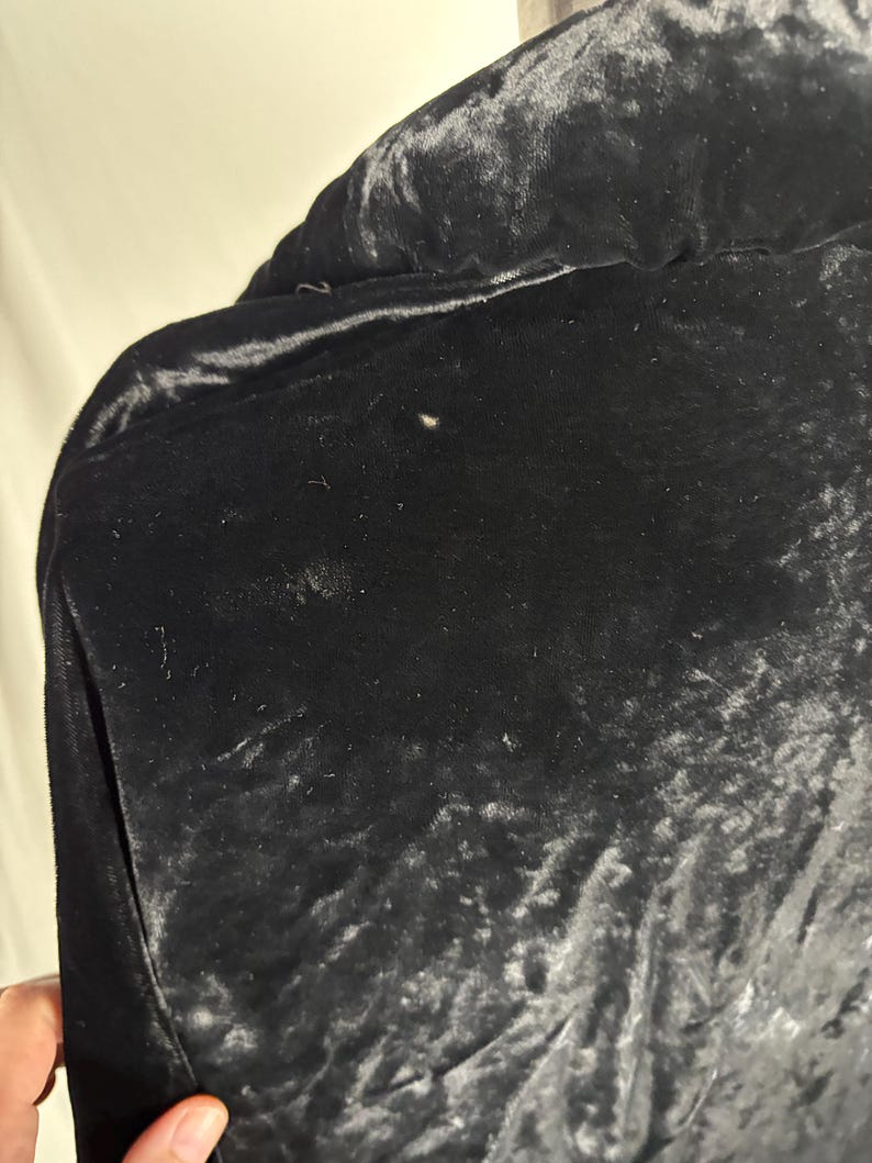 May include: Close-up of a black velvet jacket. The fabric has a soft, textured appearance with subtle variations in tone. The jacket features a pocket on the left side. The overall impression is one of luxury and elegance.