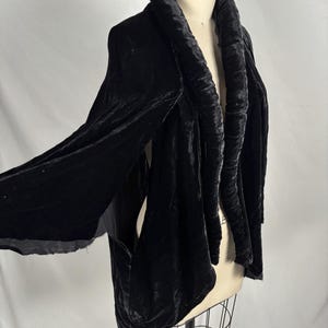 May include: A black velvet jacket with a wide, ruched collar and long sleeves. The jacket is displayed on a mannequin, showcasing its draped design and luxurious texture. The garment has a classic, elegant style.