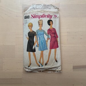 May include: Vintage Simplicity sewing pattern packet, featuring illustrations of three dresses. The packet is labeled "6849 Simplicity" and includes text about the pattern's design and sizing. The packet is slightly worn, with a price tag of 75¢.