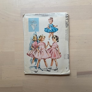 May include: Vintage McCall's sewing pattern for a child's dress and button-on pinafore. The pattern features illustrations of children's dresses in pink and blue. The pattern number is 2120, and the price is 45 cents.
