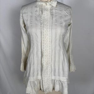 May include: A white long-sleeved blouse with vertical pin-tuck detailing and lace trim. The blouse features a ruffled collar, a lace-trimmed centre panel, and a ruffled hem. The sleeves have lace cuffs. The garment is displayed on a mannequin.