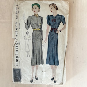 May include: A vintage Simplicity pattern envelope, number 2574, size 18, bust 36. The illustration features two dress designs: one in grey with a green hat and yellow belt, and one in blue with dark red bows. The paper envelope shows signs of wear.