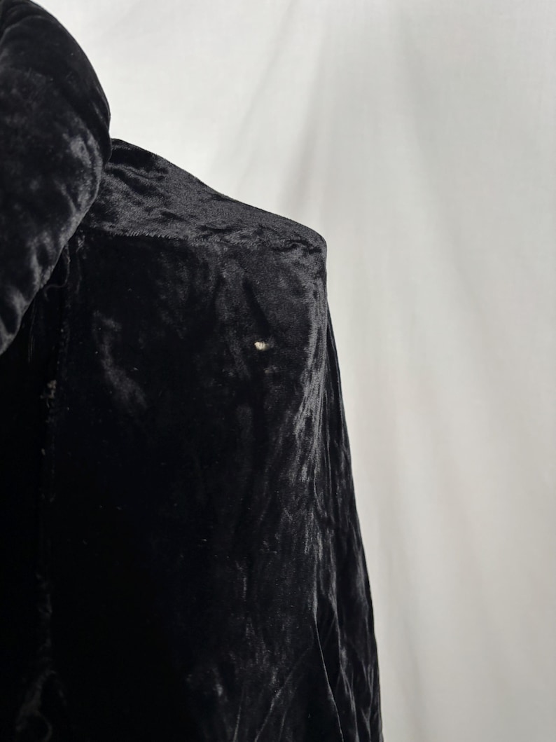 May include: Close-up of a black velvet garment, possibly a coat or jacket, with a soft, textured appearance. The fabric drapes and folds, with a small, light-colored spot visible on the surface. The garment is against a white background.