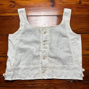 May include: A vintage, off-white linen vest with a square neckline and button-up front. The vest has shoulder straps and small buttons at the bottom hem. The garment is displayed on a wooden surface.