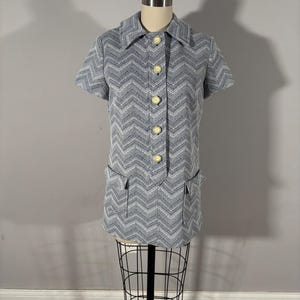 May include: A vintage short-sleeved top in a blue and white chevron pattern. The top has a collared neckline, button-down front, and two front pockets. It is displayed on a mannequin.