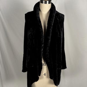 May include: A black velvet jacket with a shawl collar and long sleeves. The jacket is displayed on a mannequin, showcasing its draped design and soft texture. The garment has a flowing, open front.