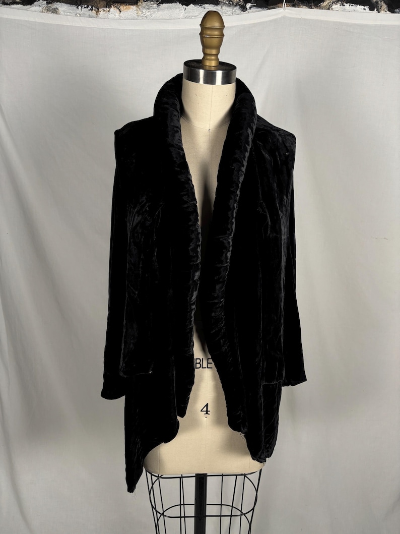 May include: A black velvet jacket with a shawl collar and long sleeves. The jacket is displayed on a mannequin, showcasing its draped design and soft texture. The garment has a flowing, open front.