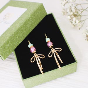May include: A pair of gold earrings with pink and light blue beads and a bow design. The earrings are displayed in a green box with a black velvet lining.